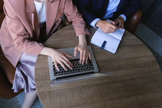 From above of crop anonymous female managers wearing formal outfits sitting at wooden table and working together with laptop and planner