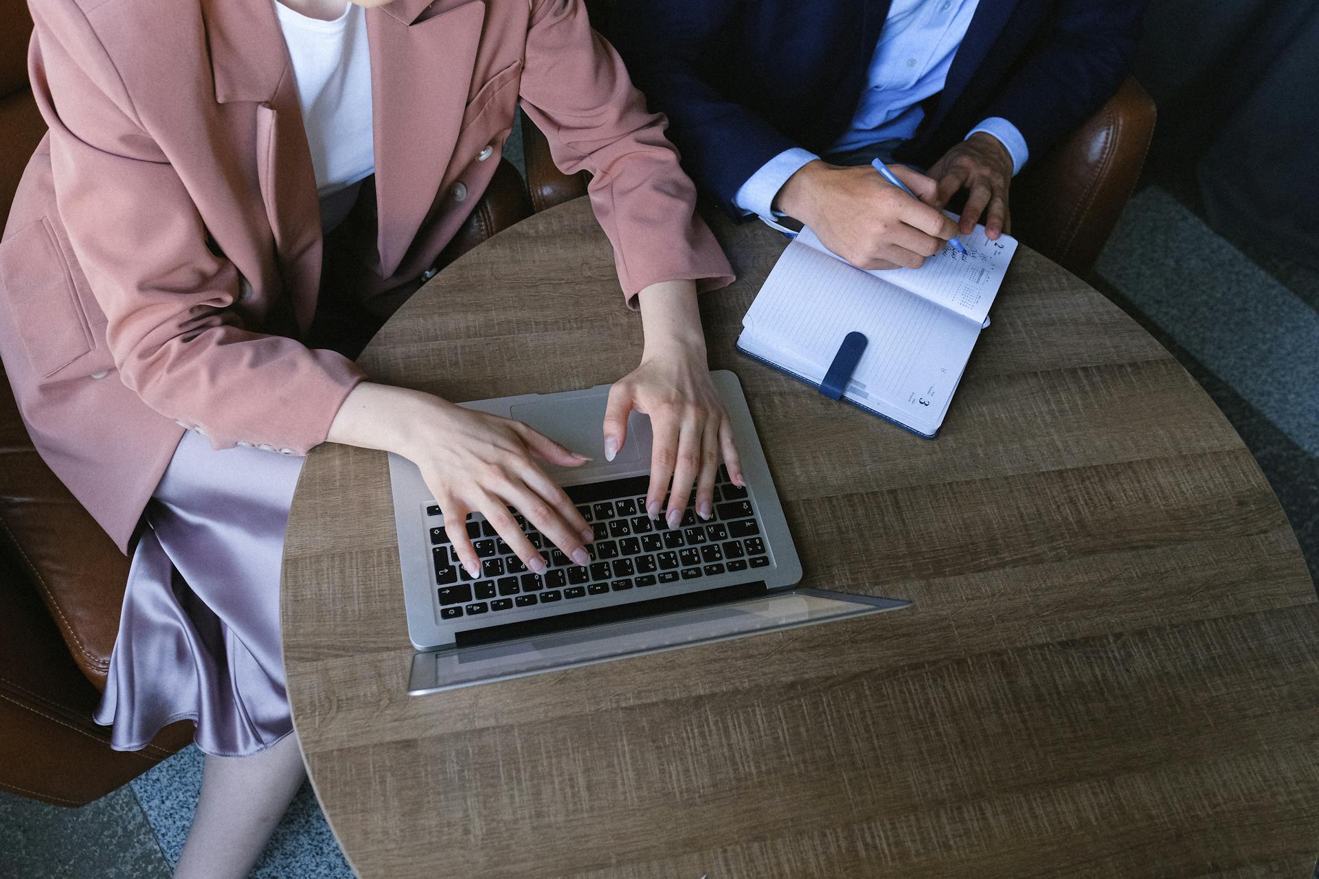 From above of crop anonymous female managers wearing formal outfits sitting at wooden table and working together with laptop and planner