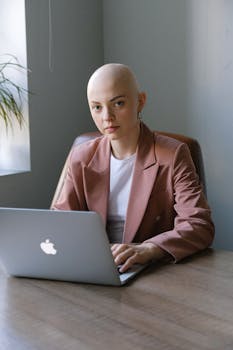 Bald woman in blazer working on laptop in bright office setting.