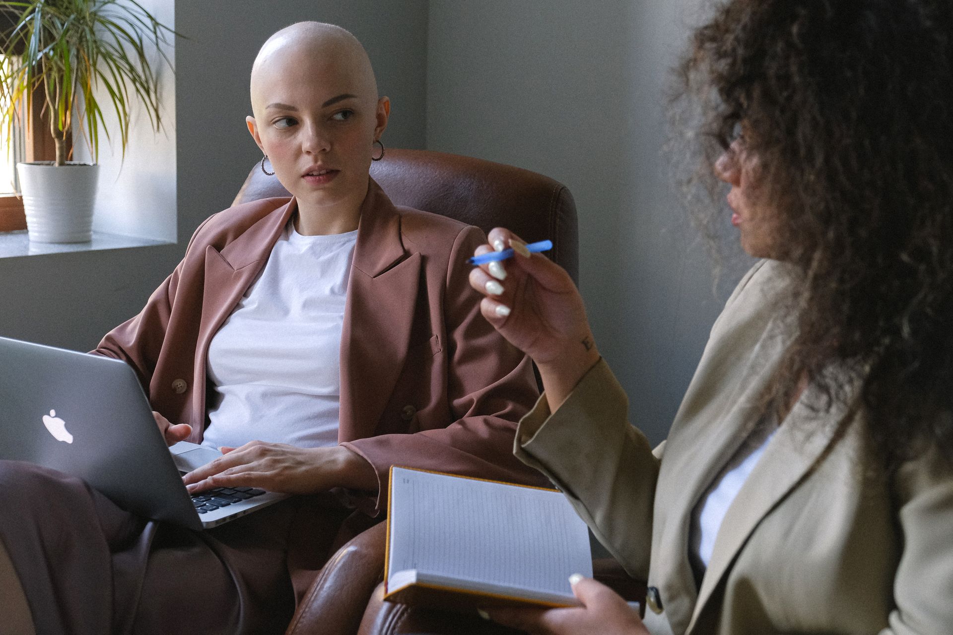 Concentrated young female entrepreneur sitting with portable laptop on knees and carefully listening to African American coworker with pen and organizer