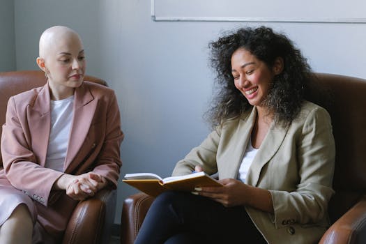 Two women enjoy a friendly conversation indoors, smiling and sharing a notebook, fostering a warm and cheerful atmosphere.