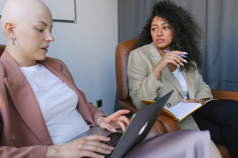 Young bald woman in stylish outfit sitting in armchair and browsing laptop while talking to female colleague with notebook