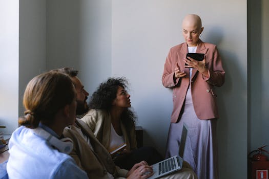 Young bald woman in trendy clothes standing near wall and browsing tablet while having conversation with colleagues sitting on couch