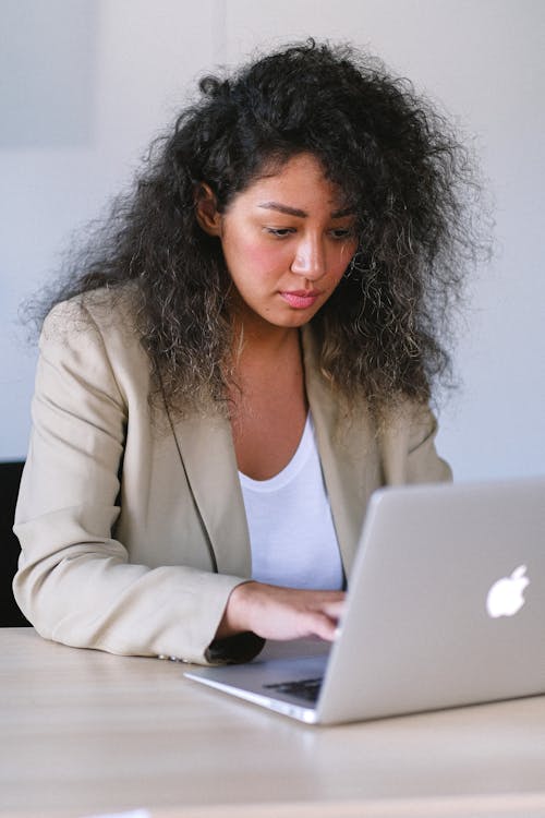 Free Young ethnic female in stylish outfit sitting at table and browsing laptop while working in office Stock Photo
