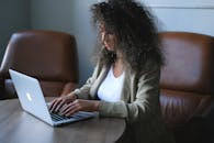 一位穿着时尚的年轻少数族裔女性坐在桌旁，侧对着镜头，在办公室里用笔记本电脑处理项目