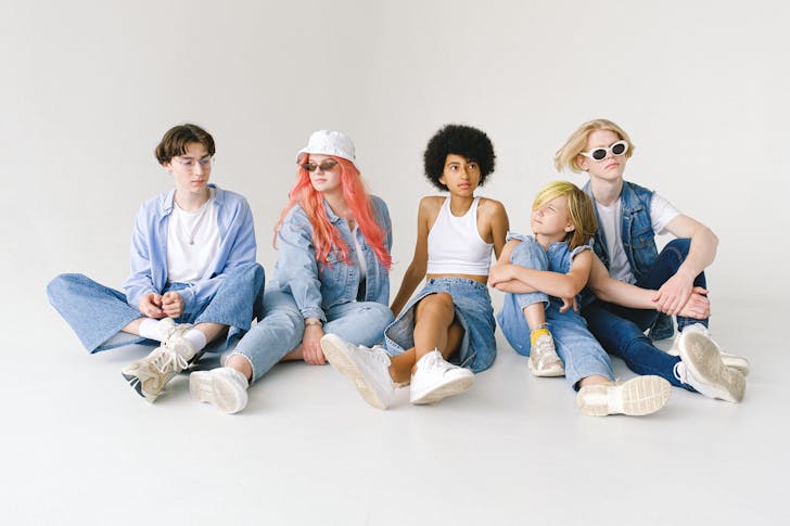 Full body of serious multiracial teens and small girl wearing denim outfits sitting on floor against white wall