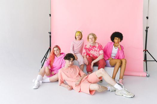 Full body informal multiracial teenager friends in pink clothes sitting near pink backdrop and looking at camera