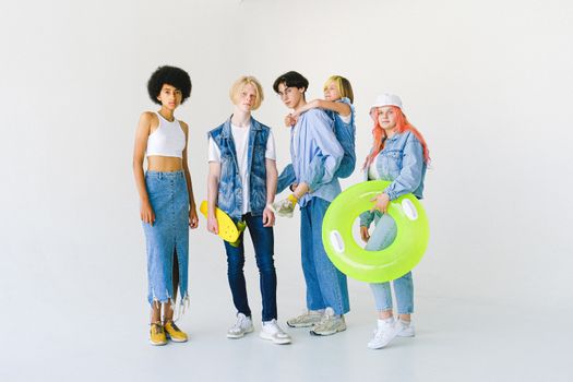 Full body of cool multiethnic teenagers and schoolchild in casual denim outfit standing in studio with inflatable ring and skateboard giving piggyback while looking at camera on white background