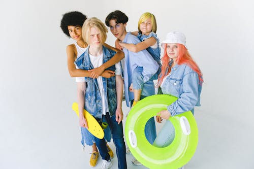 Free High angle of cool multiracial friends in casual clothes standing in studio with inflatable ring and longboard while giving piggyback ride and hugging while looking at camera on white background Stock Photo