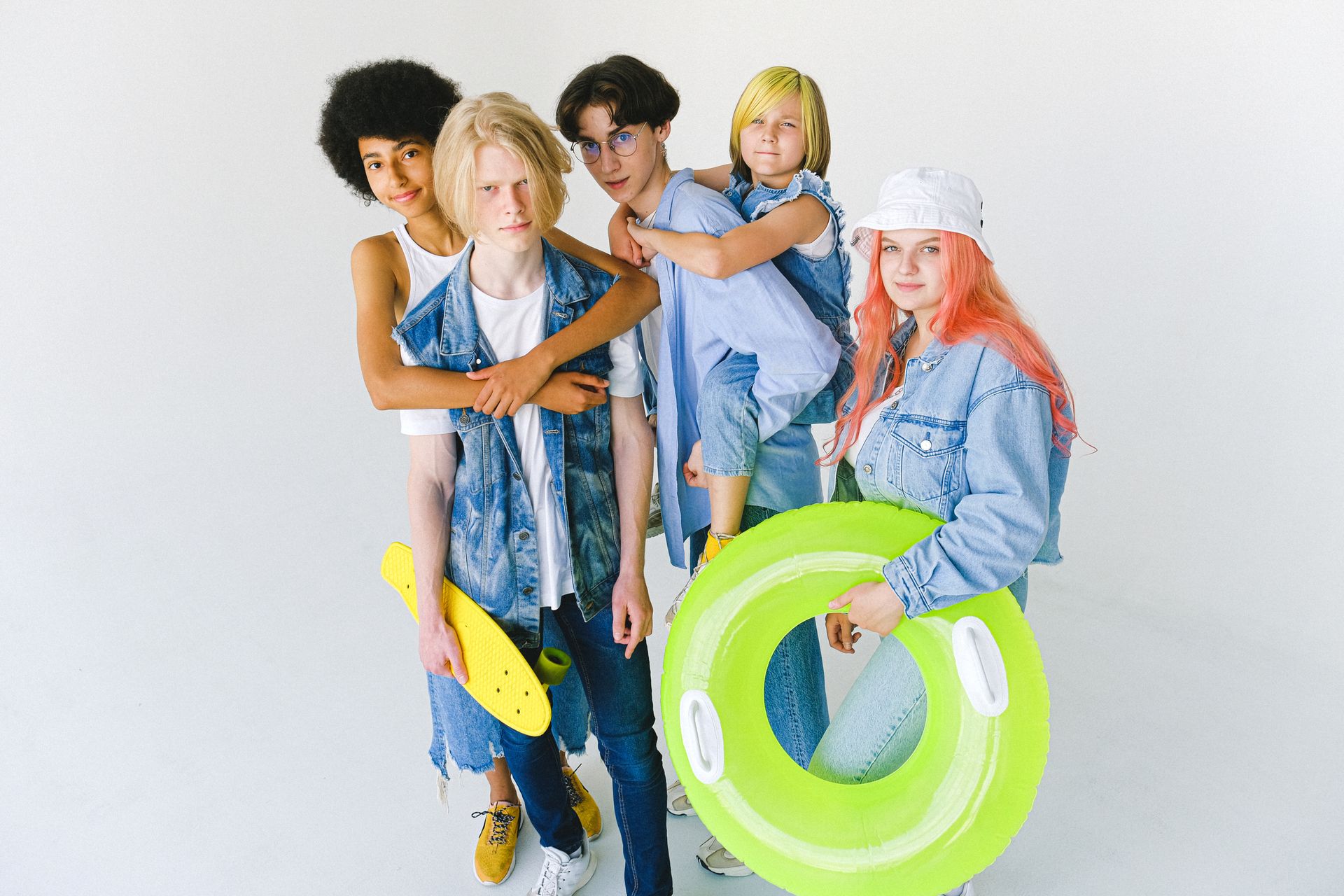 High angle of cool multiracial friends in casual clothes standing in studio with inflatable ring and longboard while giving piggyback ride and hugging while looking at camera on white background