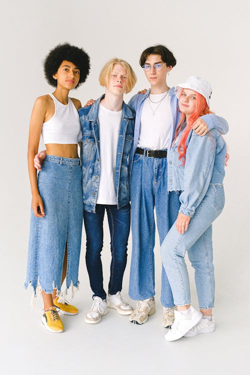 Full body of multiethnic teenagers in casual denim outfit standing in studio and hugging on white background while looking at camera