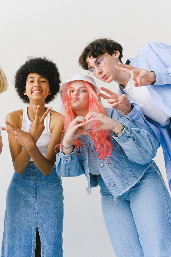 Low angle of delighted teenager friends in denim wear looking at camera while making gestures and standing against white background