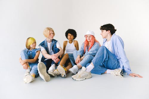 Full body group of thoughtful multiethnic teenager friends in jeans clothes sitting on floor and speaking with each other against white background