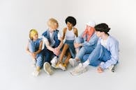 High angle of full body group of thoughtful diverse informal teenager friends sitting on floor and talking to each other against white background