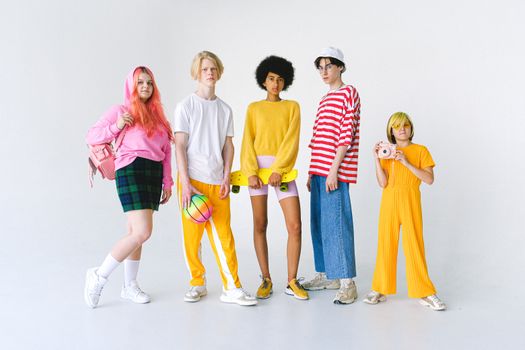 Full body group of informal teenager friends in colorful outfits standing against white background with ball and penny board with photo camera and backpack looking at camera