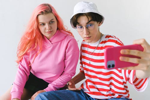From above of focused informal teenager couple in bright colorful clothes sitting and taking selfie on smartphone against white background