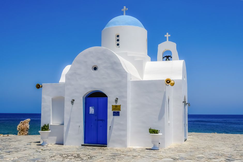 Whitewashed Cypriot Orthodox chapel with blue dome beside the sea under bright Mediterranean sun.