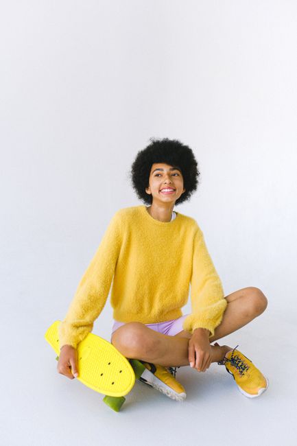 Full body happy teenage African American female skater sitting against white background with yellow penny board and legs crossed and looking away