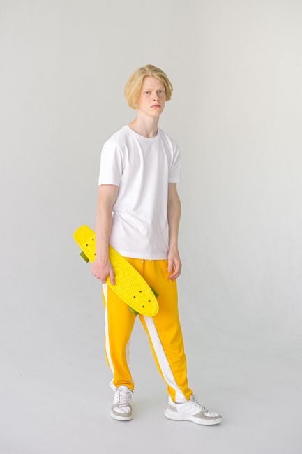 Full body of teenage boy with skateboard in colorful yellow trousers and sneakers looking at camera on white background
