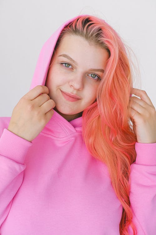 Content teen smiling and looking at camera while wearing trendy pink hoodie on white background of studio