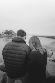 Black and white back view of unrecognizable couple in warm outerwear standing on stony shore near calm river in nature