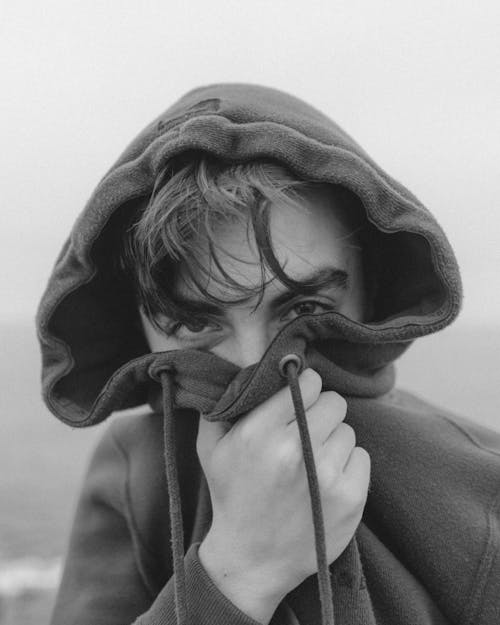 Black and white of unrecognizable young male traveler covering mouth with hood and looking at camera while relaxing on seashore on gloomy day