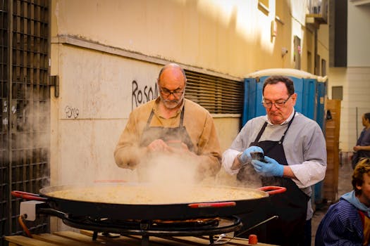 Two chefs cook a large paella outdoors using a phone recipe.
