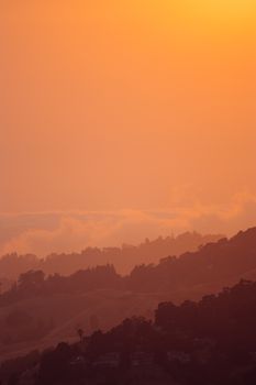 Lush dark green forest on mountain slopes under highlighted clouds and vivid orange sky at dawn