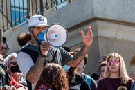 Ethnic male wearing white hat standing near concrete stair with loudspeaker near crowd of people during protest on street in summer day