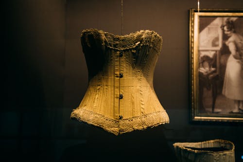 Bright historical corset with buttons hanging on cord in showcase near picture in museum