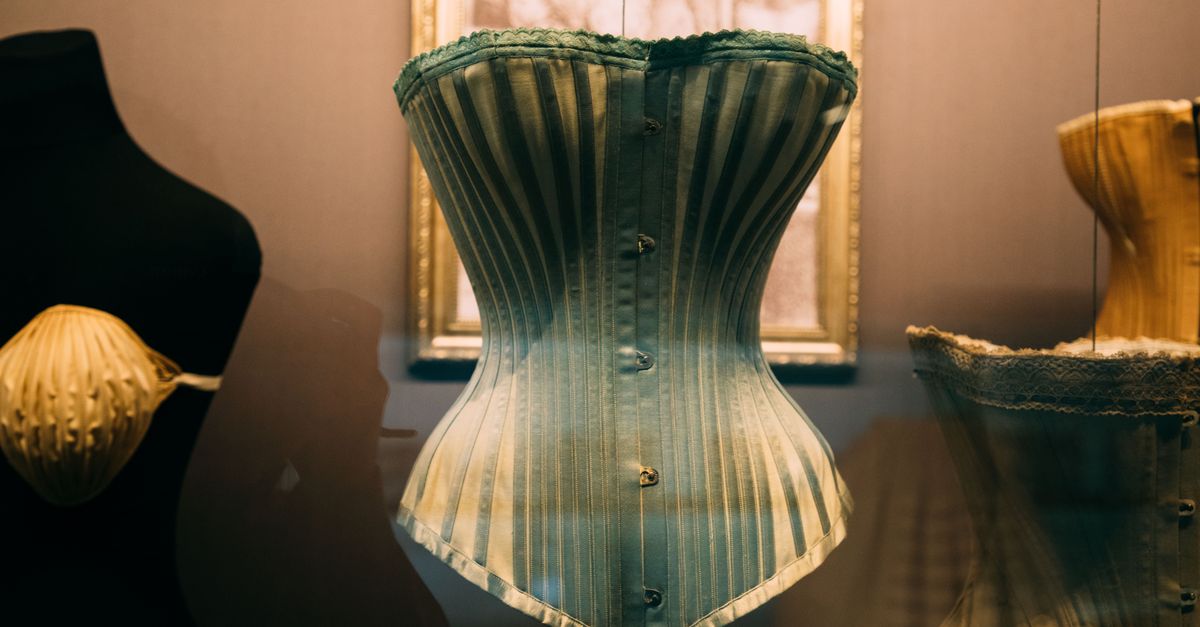 Old style feminine corset with buttons and stripes hanging on cord in showcase near picture on wall