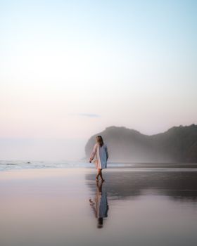 日落时分，一名女子沿着宁静的海滩散步，在潮湿的沙滩上投下宁静的倒影