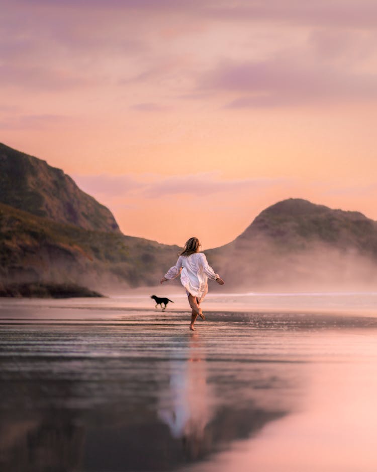 Unrecognizable Woman With Dog Running On Picturesque Beach In Twilight