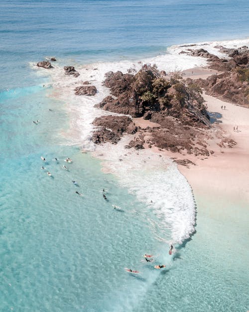 Drone view of anonymous travelers swimming in pure rippled sea near sandy beach with rocks in summer