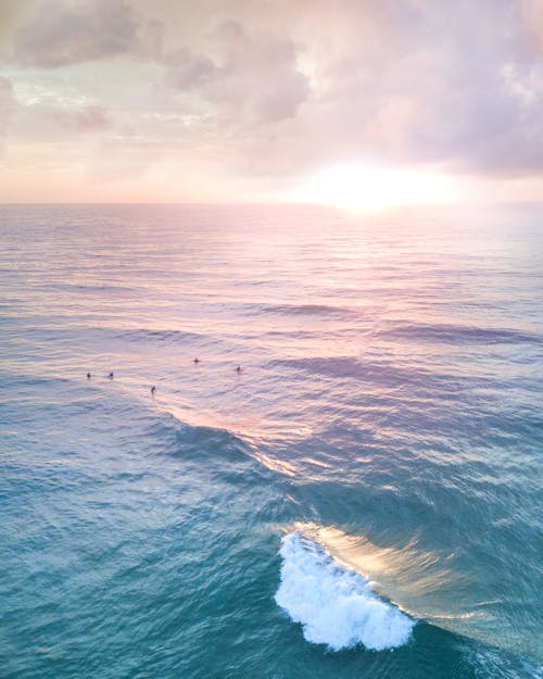 From above of anonymous travelers swimming in rippled sea with foam under shiny cloudy sky in evening