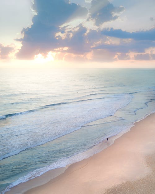 From above of unrecognizable tourist on sandy ocean coast under cloudy sky with shiny sunlight in evening