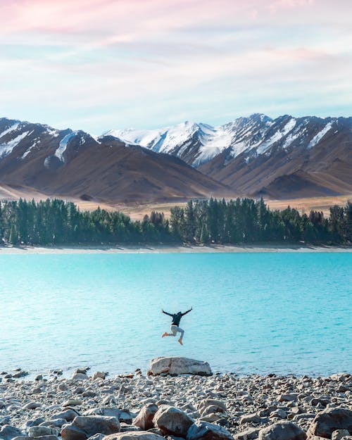 Back view of unrecognizable person jumping on rocky coast near calm turquoise river against mountains with snow in daytime