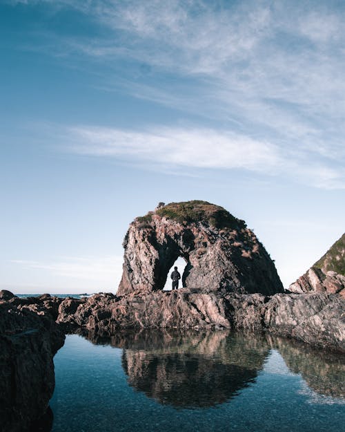 Back view of anonymous person standing in rocky arch near calm water against blue sky
