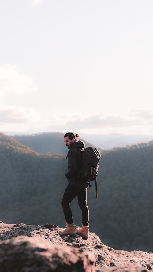 Side view of man in warm clothes carrying backpack standing on edge of rocky mountain in sunny day