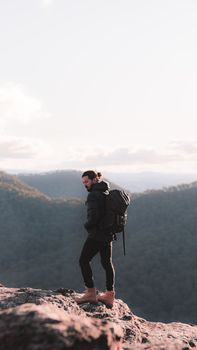 Side view of man in warm clothes carrying backpack standing on edge of rocky mountain in sunny day