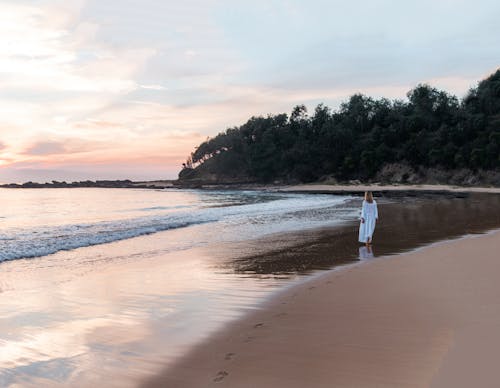 Back view of anonymous female in long white dress walking along empty sandy beach near calm ocean at sunset