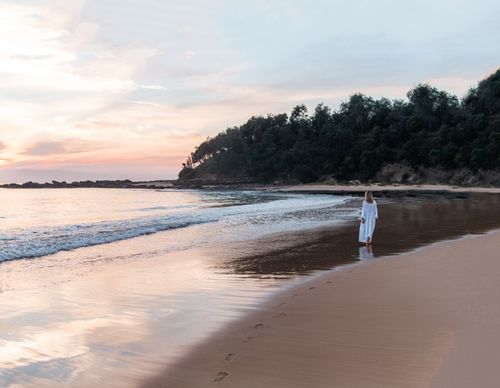 Free Back view of anonymous female in long white dress walking along empty sandy beach near calm ocean at sunset Stock Photo