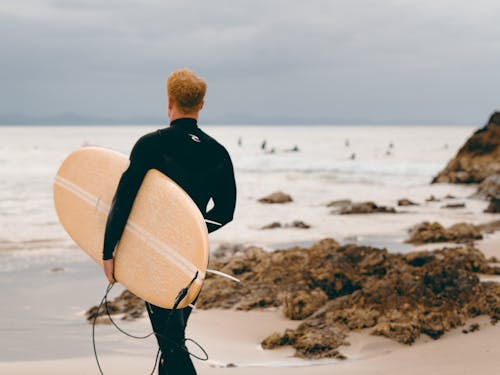해변에 베이지 색과 흰색 서핑 보드를 들고 검은 재킷을 입은 남자
