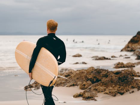 Surfer in wetsuit holding board, facing the ocean at Byron Bay, Australia.