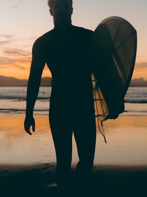 Schattenbild Des Mannes, Der Surfbrett Am Strand Während Des Sonnenuntergangs Hält