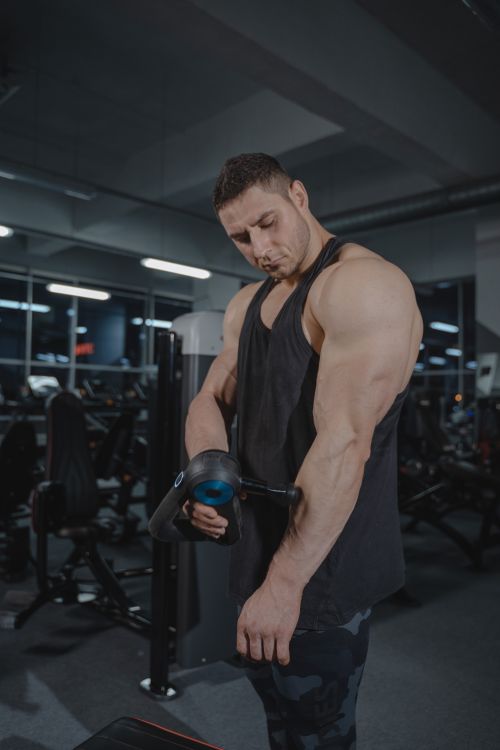 Free A muscular man uses a massage gun in a gym, showcasing fitness and recovery. Stock Photo