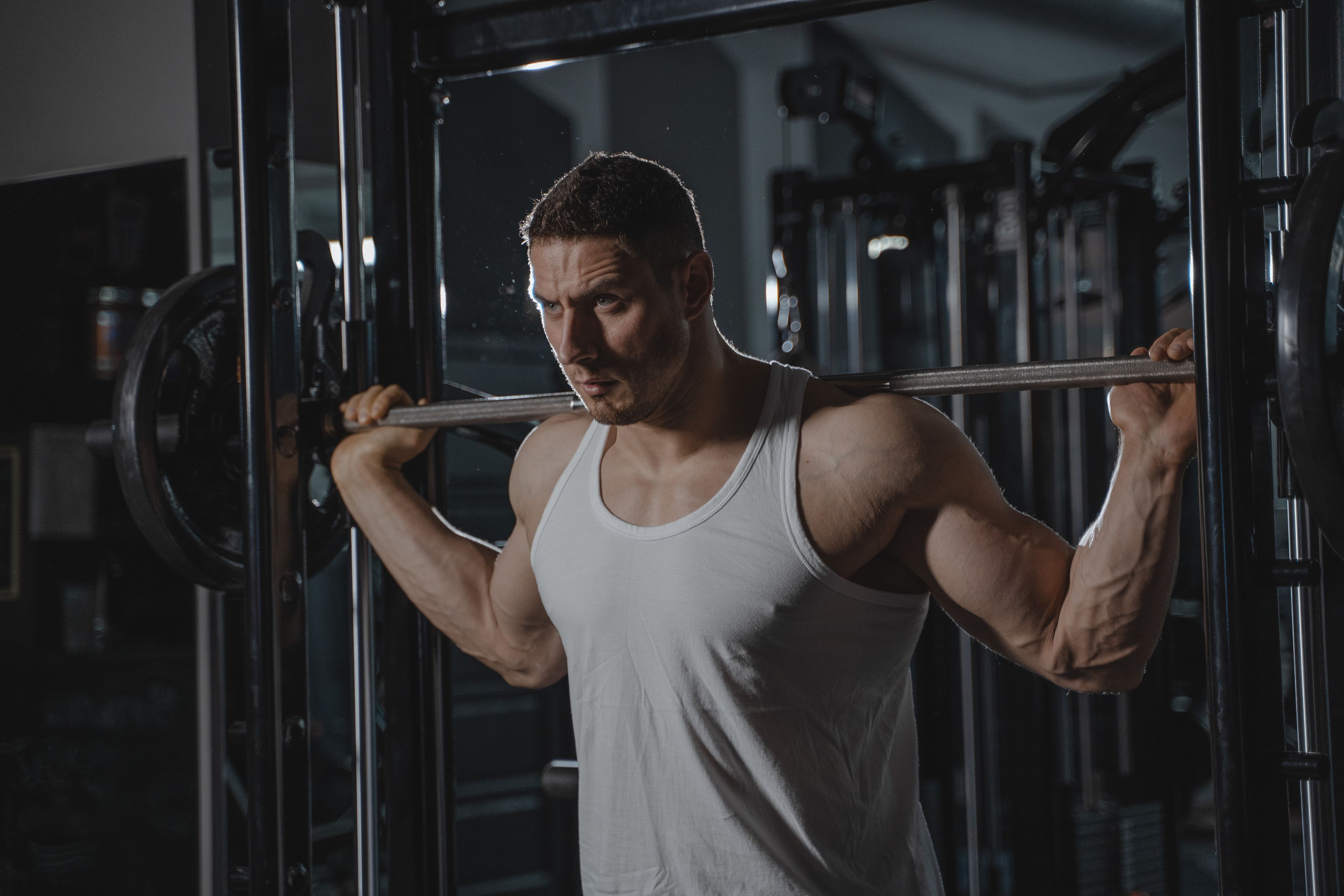 Strong man lifting weights at gym wearing a white tank top, showcasing muscle strength and fitness.