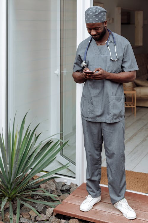 A Nurse in Gray Scrubs Suit Standing Beside the Glass Door while Busy Using His Cellphone
