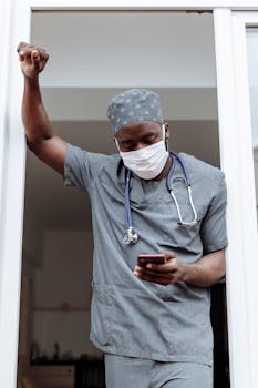 A healthcare worker in scrubs and face mask checking phone in doorway.