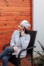 一位穿着医用手术服的女人微笑着坐在户外木墙旁打电话